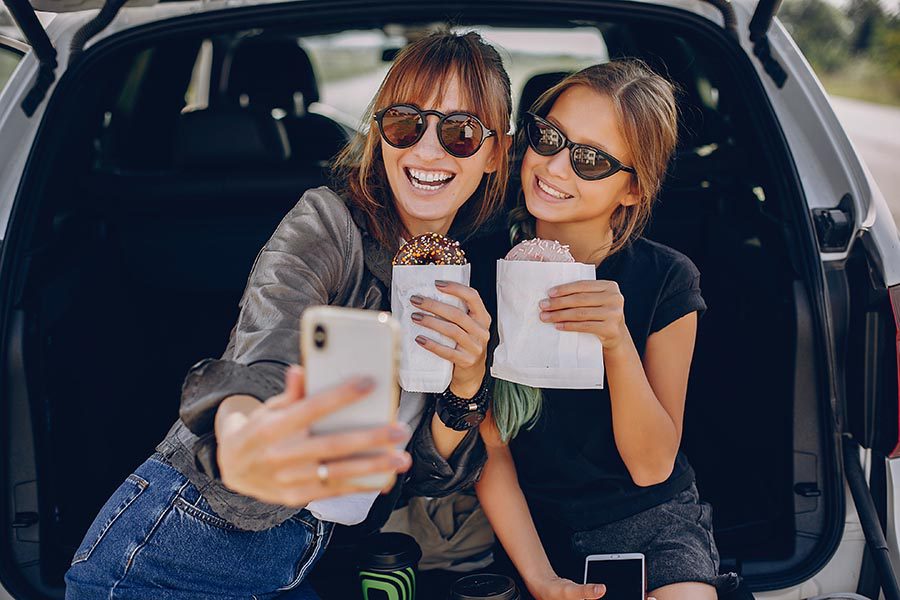 Personal Insurance - Mom and Daughter Take a Selfie in the Back of Their Car, Holding Doughnuts and Wearing Sunglasses